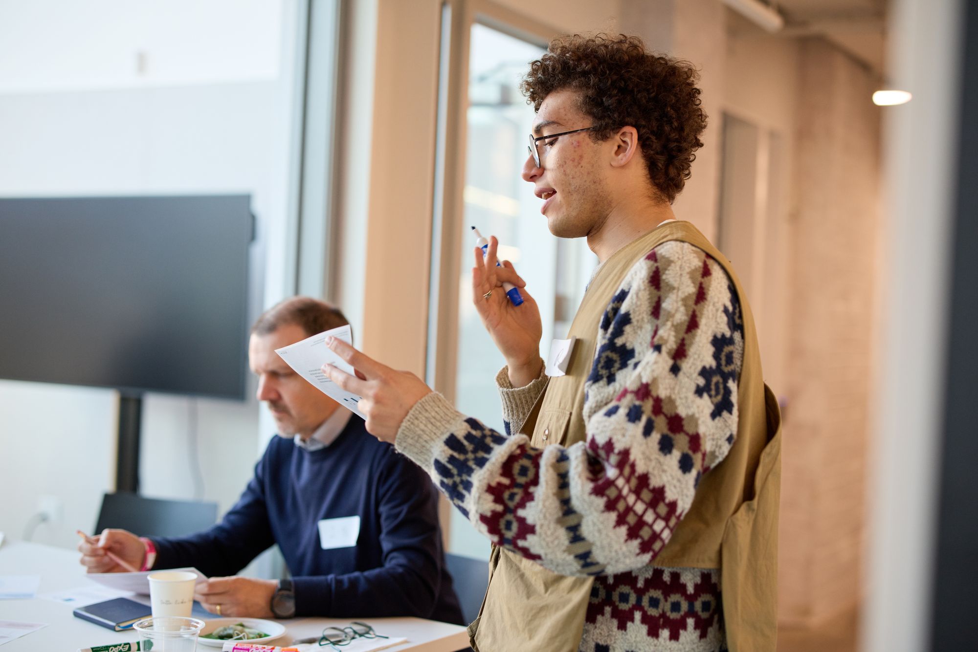 A participant in a patterned sweater speaks, gesturing with a whiteboard marker.